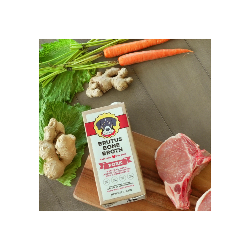 Pork Brutus Bone Broth box lying on a cutting board with raw carrots, ginger, lettuce, pork, and potatoes around it.